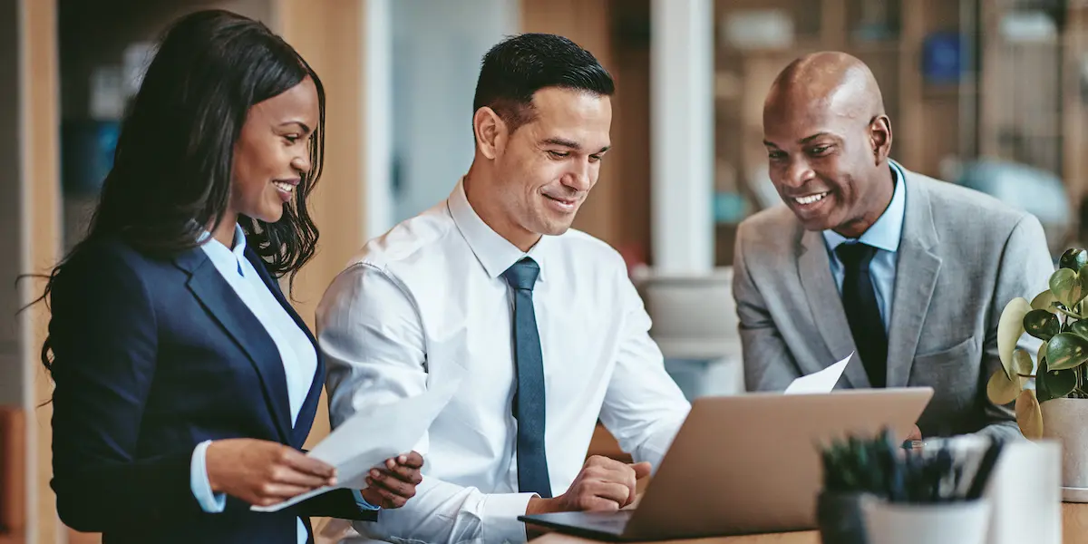 Corporate image of a group of people in suits looking at a laptop and smiling at what they see.