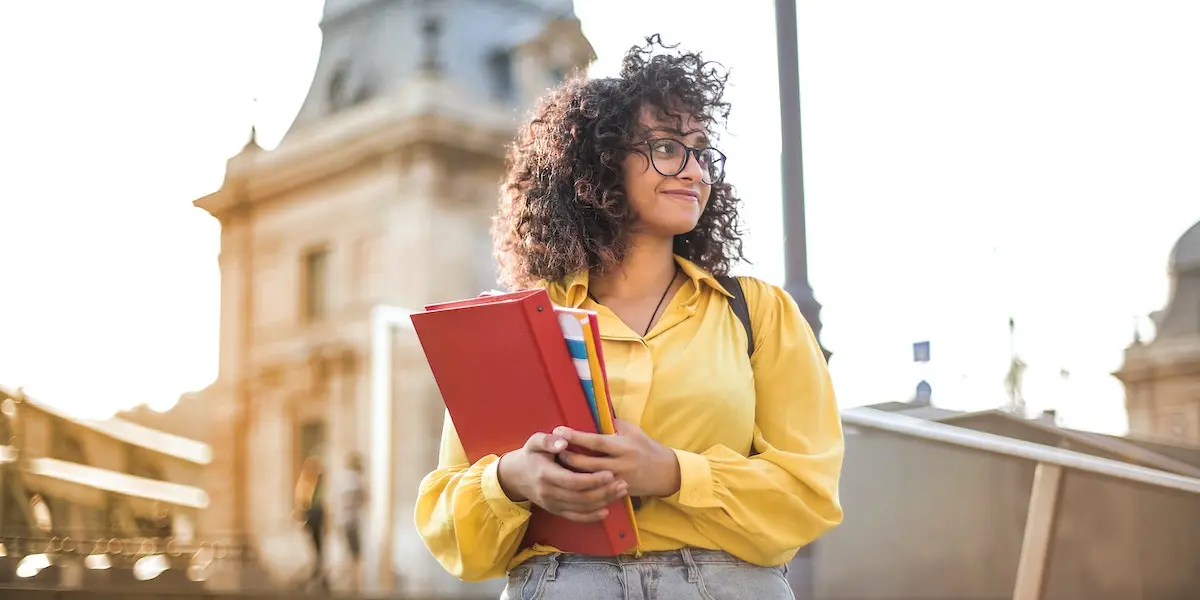 Education image of a young woman holding a notebook standing in front of a college building.