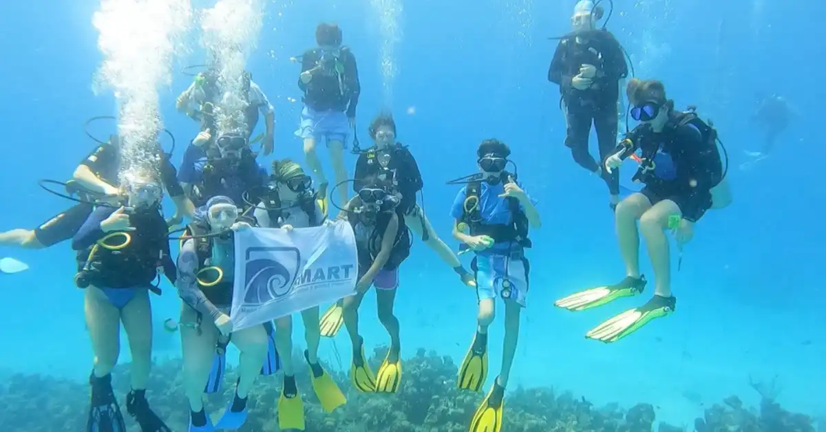 Youth Scuba divers on the sea floor, holding a SeaSmart flag.