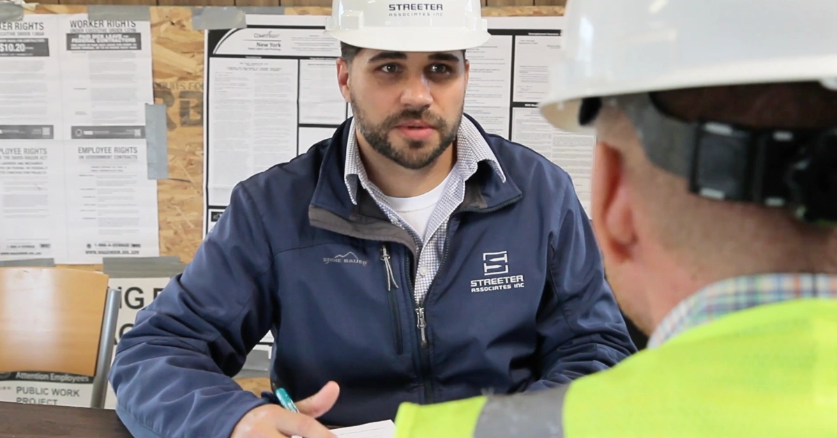 A construction worker wearing a safety hat talks with another employee in a room with worker rights posters.